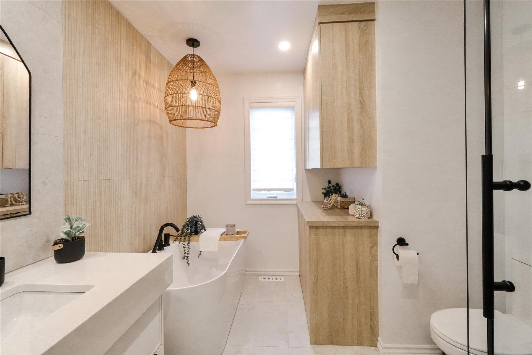 L’Oasis : Bain autoportant sous une fenêtre, vanité et armoires en bois clair, dans une ambiance chaleureuse