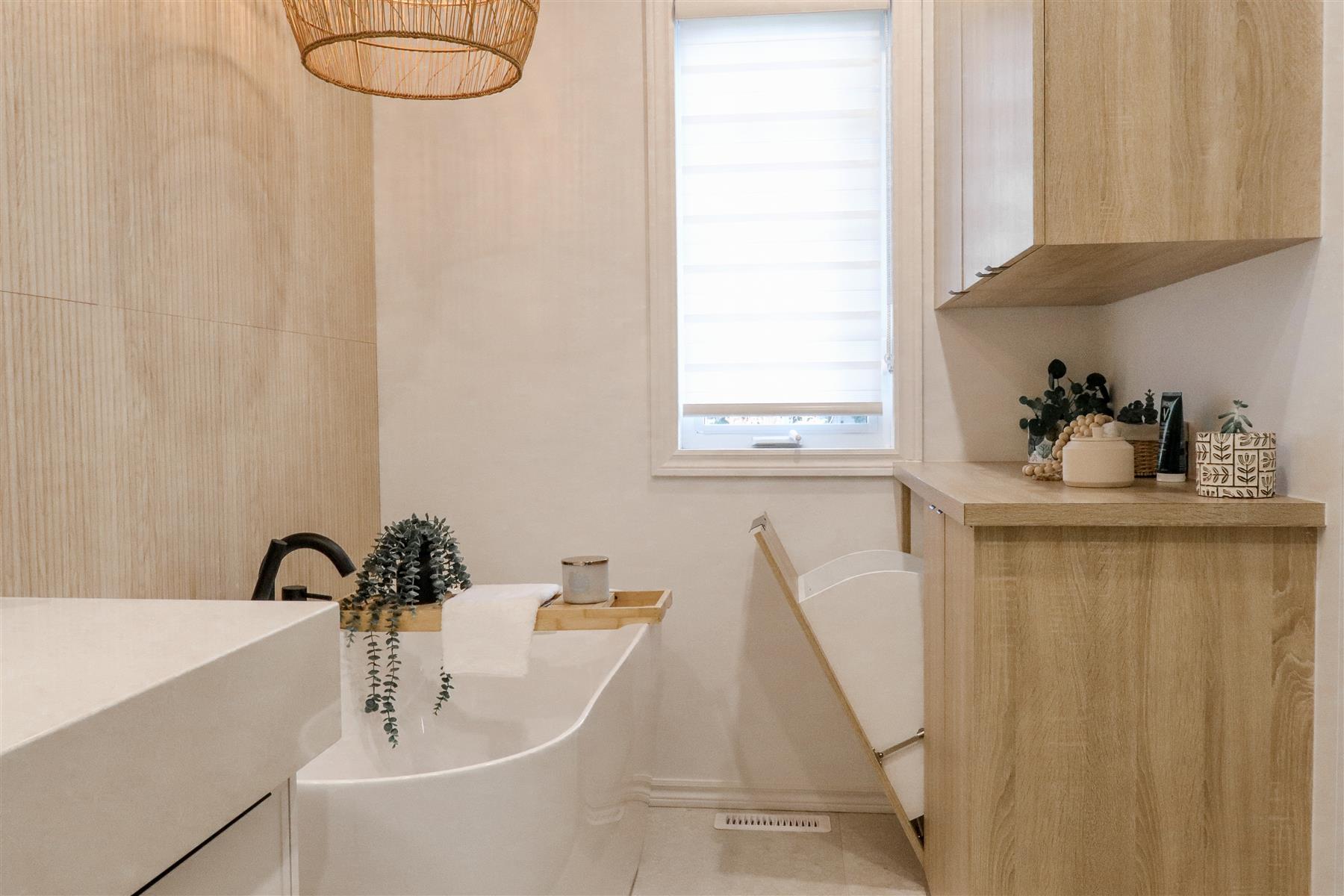 L’Oasis : Bain autoportant sous une fenêtre, vanité et armoires en bois clair, dans une ambiance chaleureuse